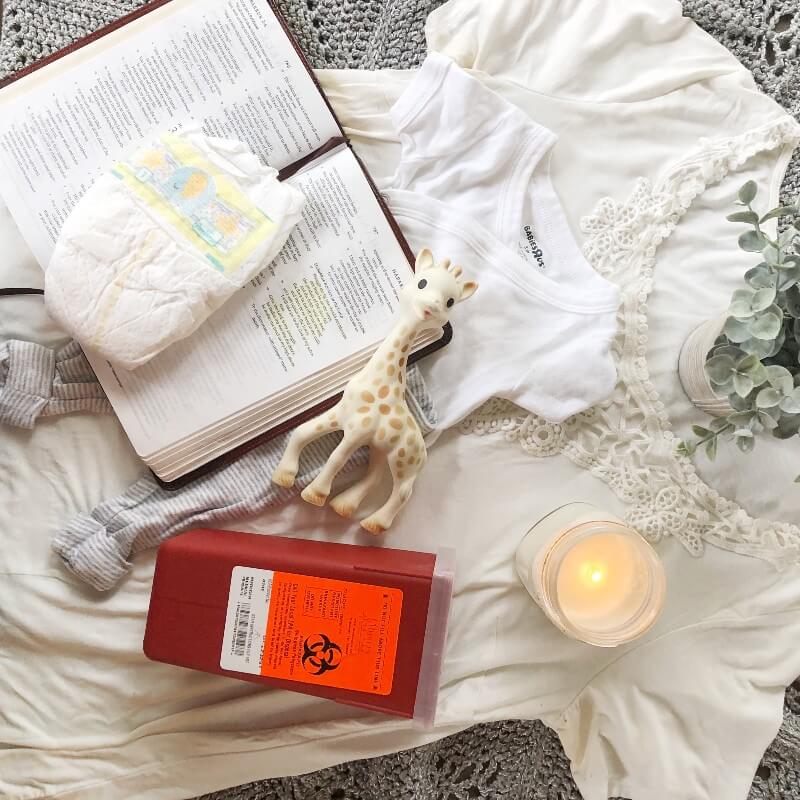 A flat lay of a maternity shirt, baby outfit, diaper, bible, teething giraffe, lit candle, and a sharps container that holds infertility injectable medications.