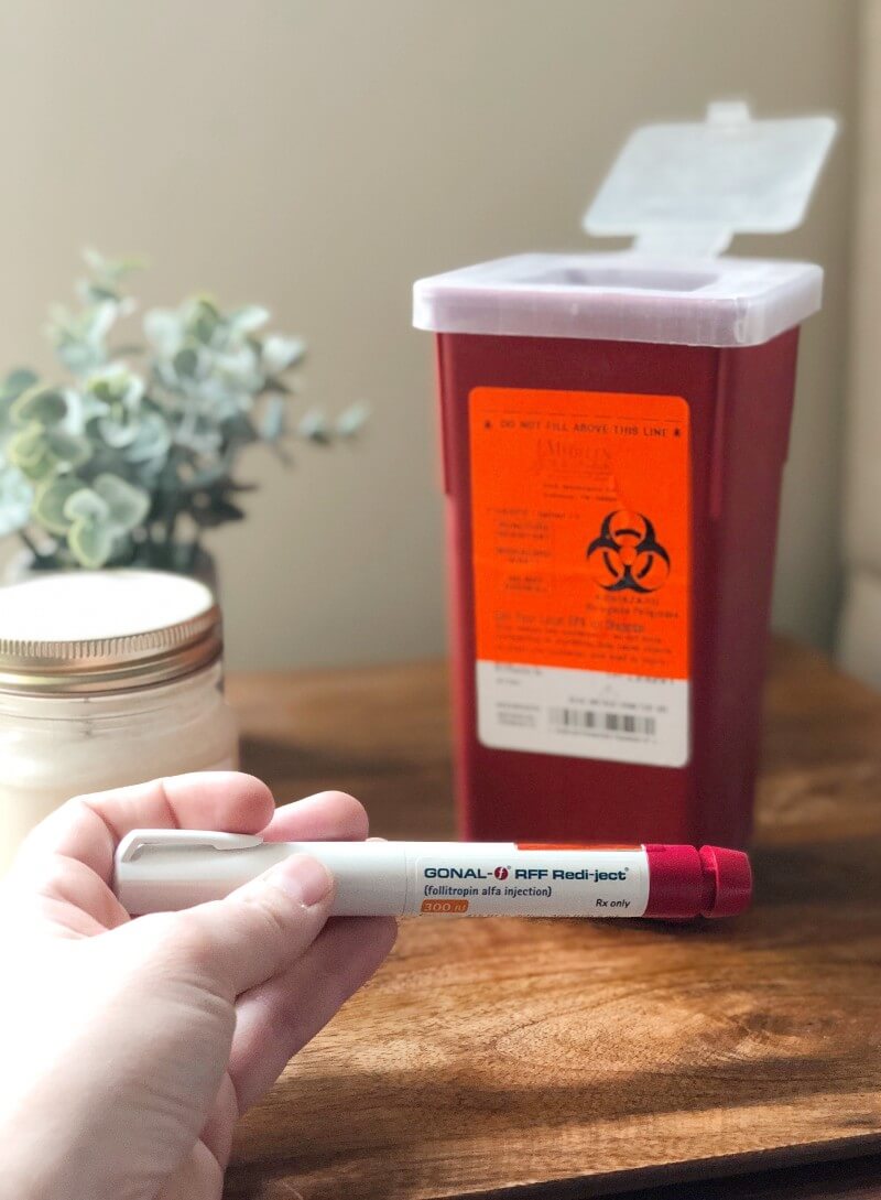 A woman's hand holding a Gonal-F injectable medication pen in front of a sharps container.