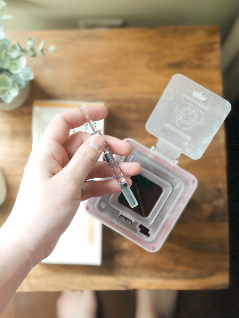 A woman's hand holding a syringe of infertility medication over the open top of a sharps containers.