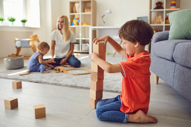 Mom sitting in living room with two playing children. Older boy is playing with blocks and baby is playing a wooden train set.