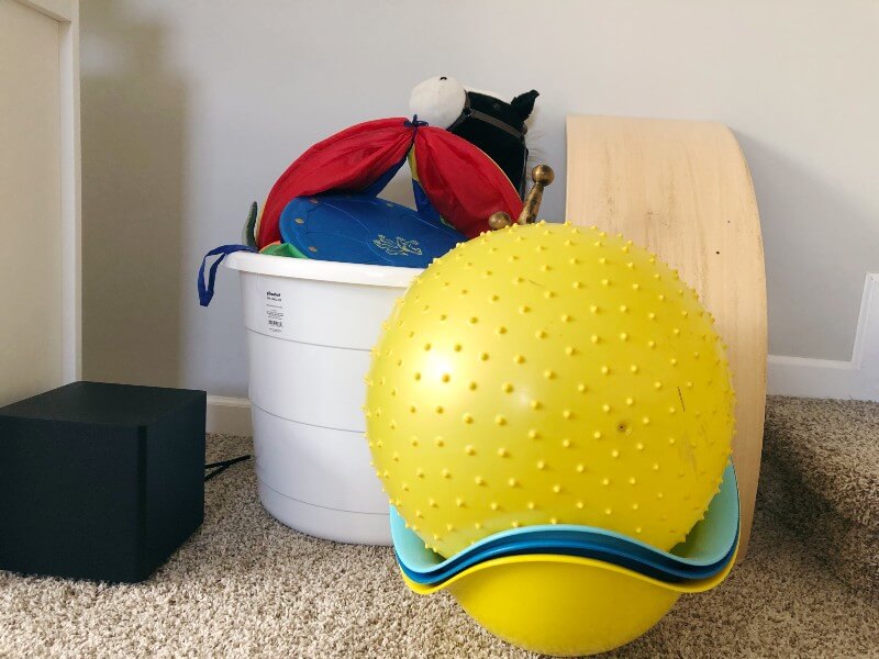Large bucket with pretend play toys inside, and large ball, wobble board, and Bilibo open ended play toys stacked next to it.