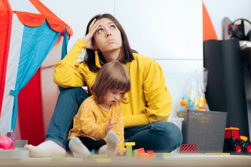 Frustrated mom sitting near playing child in a bunch of toys.